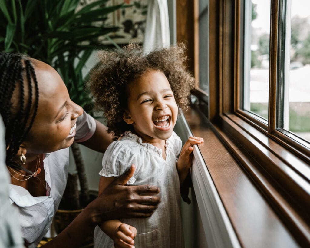 A heartwarming scene inside a Montreal home, capturing a mother and her young daughter laughing together by a window. The child, with her curly hair bouncing, flashes a bright, infectious smile while her mother, with braided hair, gently holds her, both basking in a moment of pure joy and light