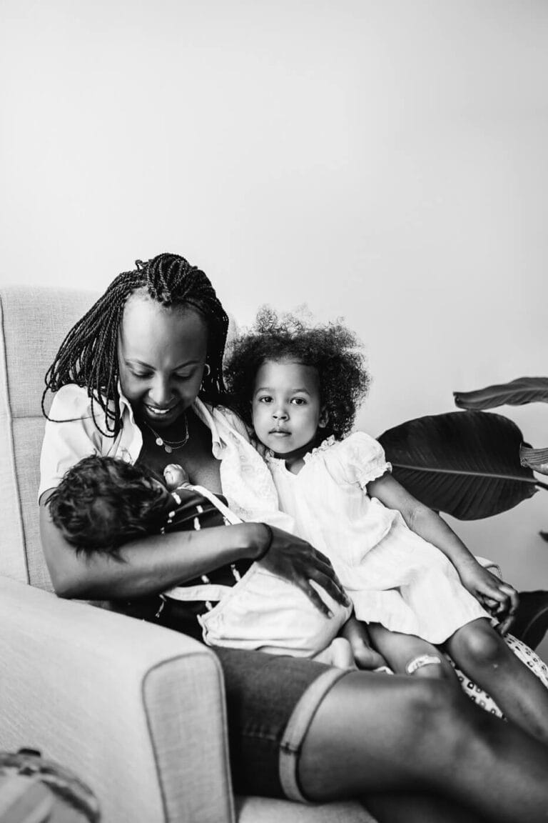 Black & White image of woman breastfeeding baby with little girl sitting on her lap looking at camera