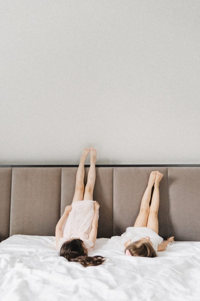 Two children lying on a bed with their legs raised against a neutral-toned headboard, wearing light dresses, in a candid, peaceful moment of sibling relaxation at home.