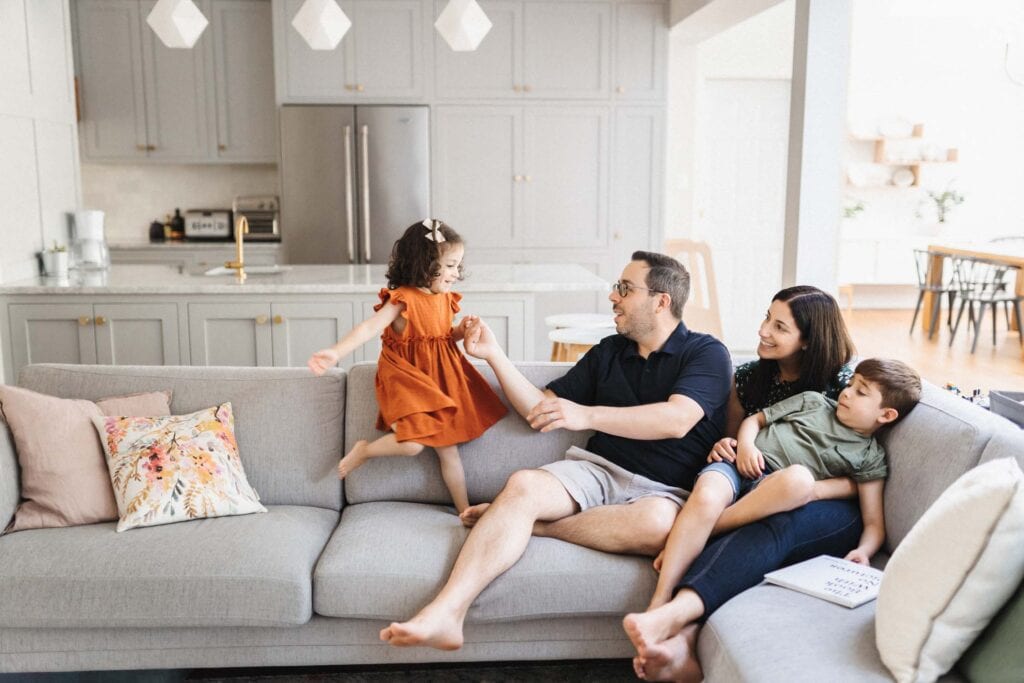 A vibrant family moment inside a stylish modern living room, featuring a father lifting his young daughter in a playful twirl, with the mother and son sitting beside them, all enjoying their time together in a cozy, well-lit space.