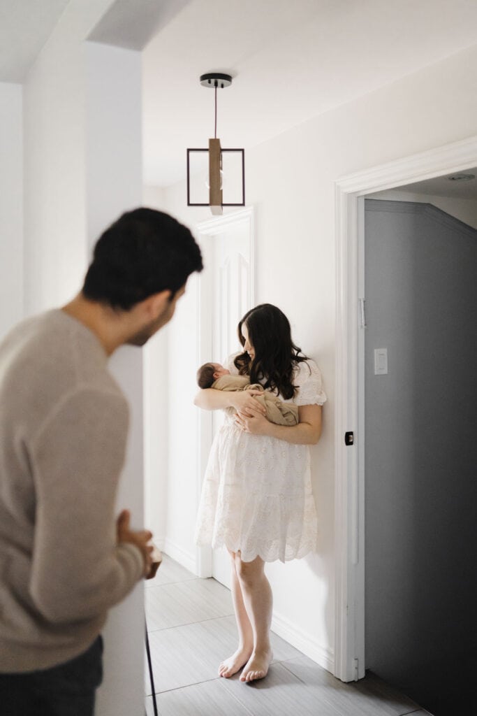 woman holds newborn baby in hallway while dad watches from behind a wall