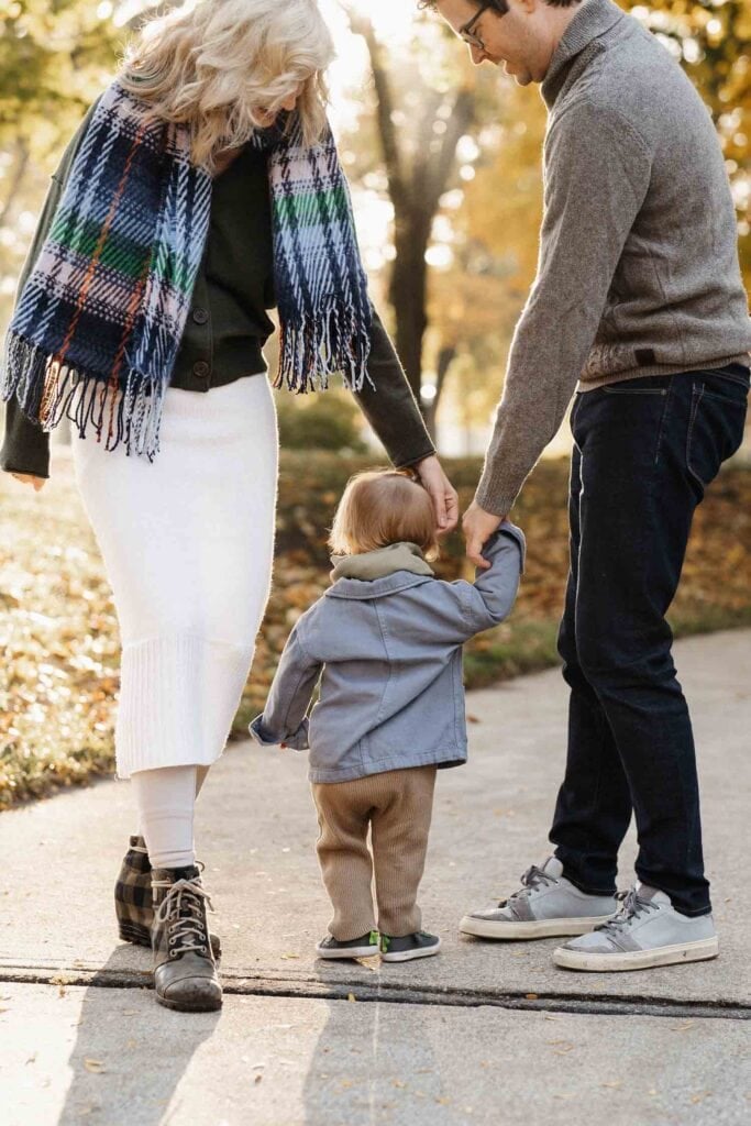 man and woman help baby walk at park in autumn