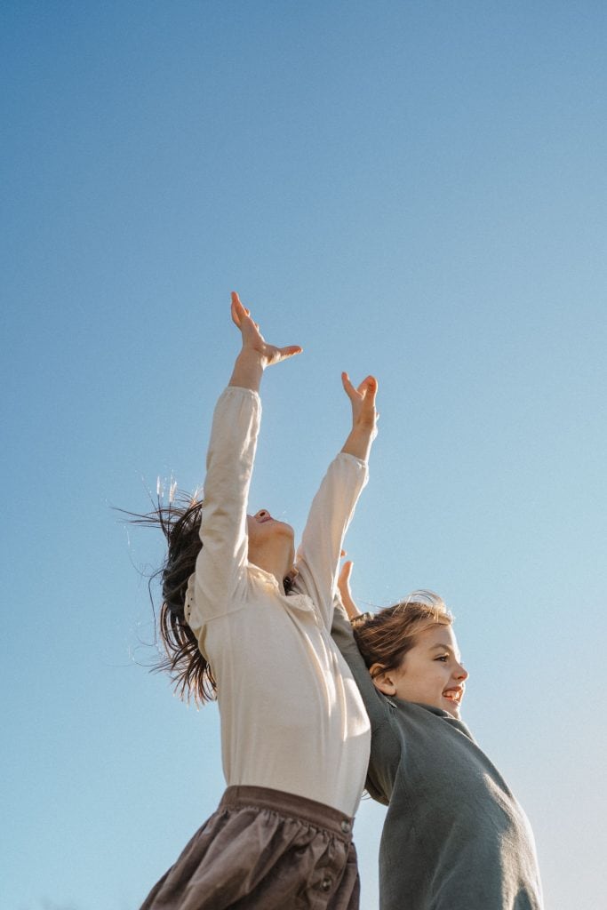 Two children reaching up to the sky in laughter, captured in a candid family photography session in Montreal. The older child in a grey sweater and the younger in a white shirt, both embody the spirit of joy under a clear blue sky.