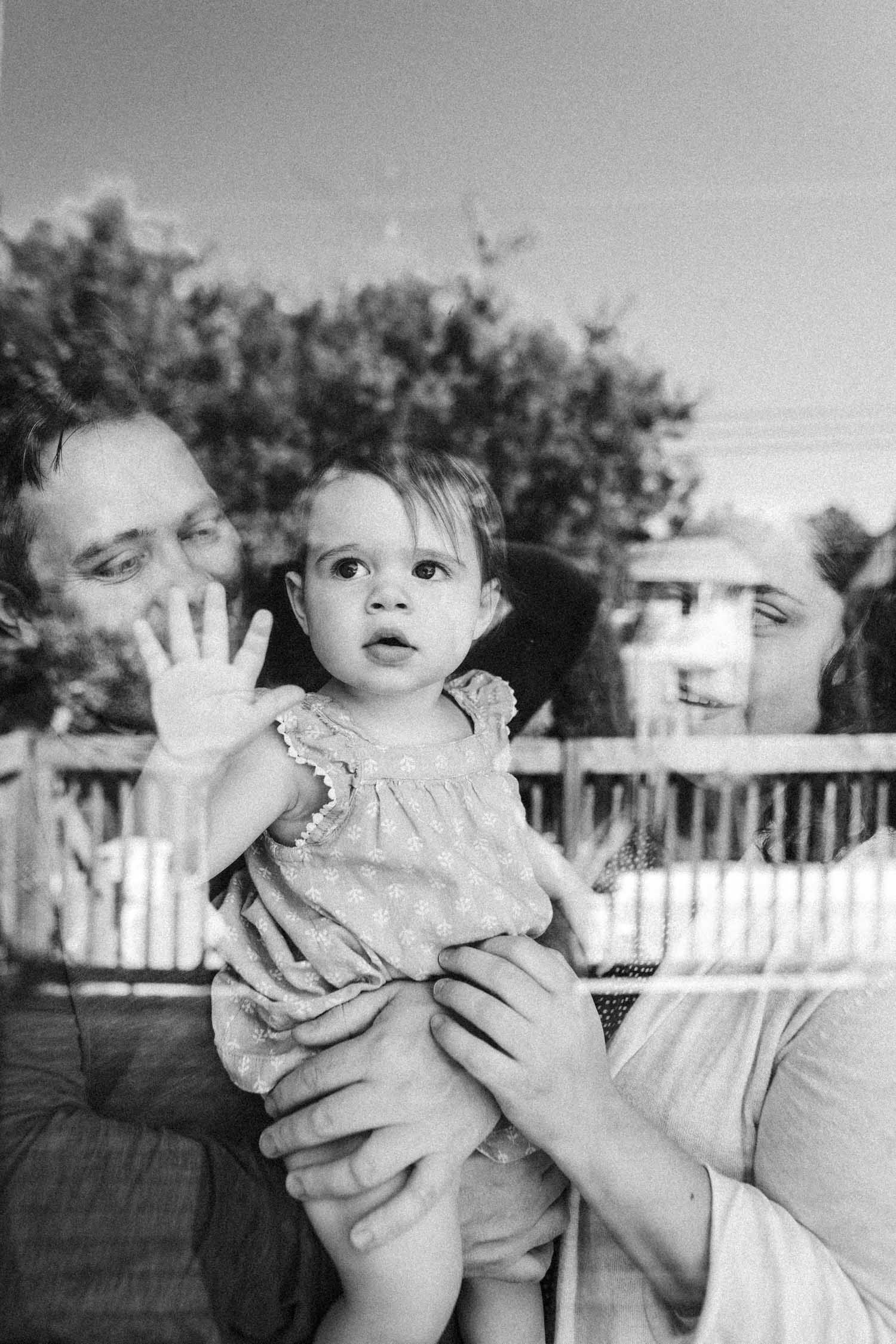A black and white photo capturing a toddler with wide eyes and an open hand, as she is held by her father, both partially visible in a charming, natural family moment. seen through a window