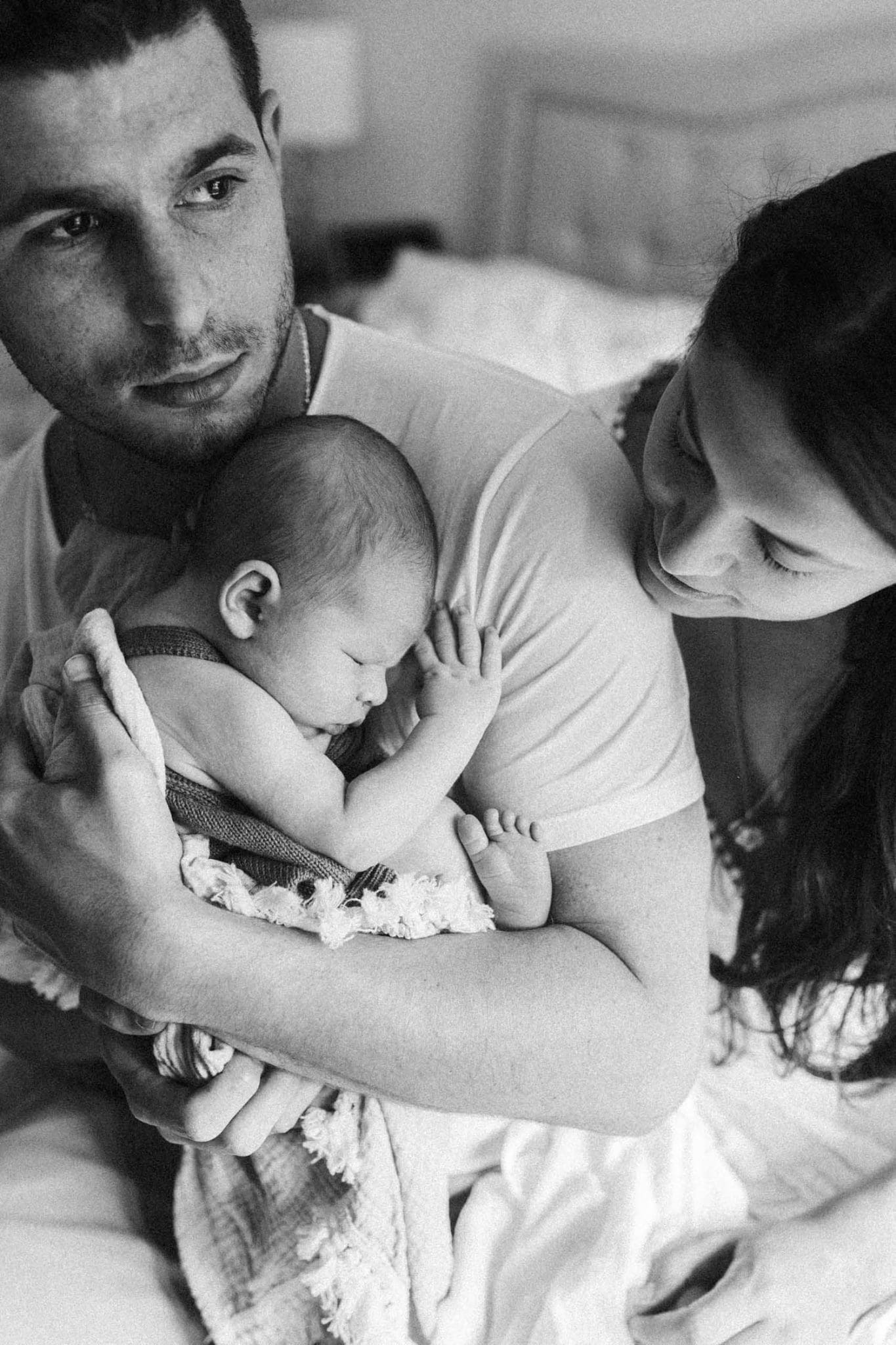 mom leans over dad's shoulder as he holds newborn baby chest to chest during in home newborn baby photo shoot in montreal