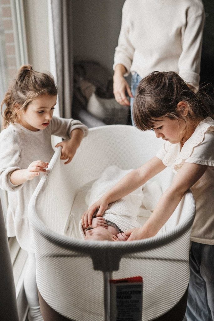 two older sisters look at their newborn baby sister in her bassinet during their in-home photoshoot in montreal