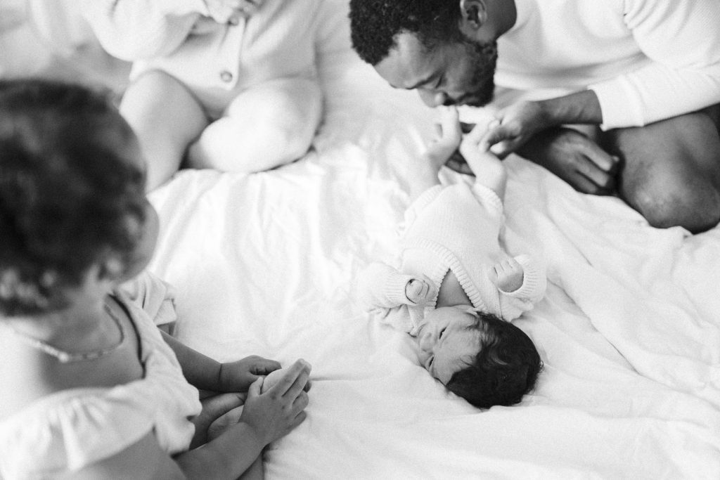 A black and white image capturing a serene moment with a Montreal family and their newborn. The father, sitting on a white bed, gently touches the baby's foot, while the mother, clad in a light outfit, watches lovingly. An older sibling, partially visible, looks on, completing this intimate family scene.
