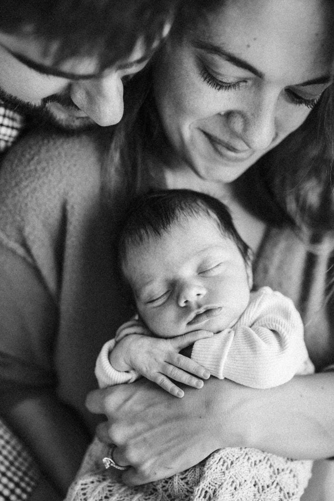 A black and white image of a young couple holding their sleeping newborn close, capturing a moment of profound tenderness. The parents, gently embracing their child, showcase the raw, emotional essence characteristic of candid newborn photography in Montreal.