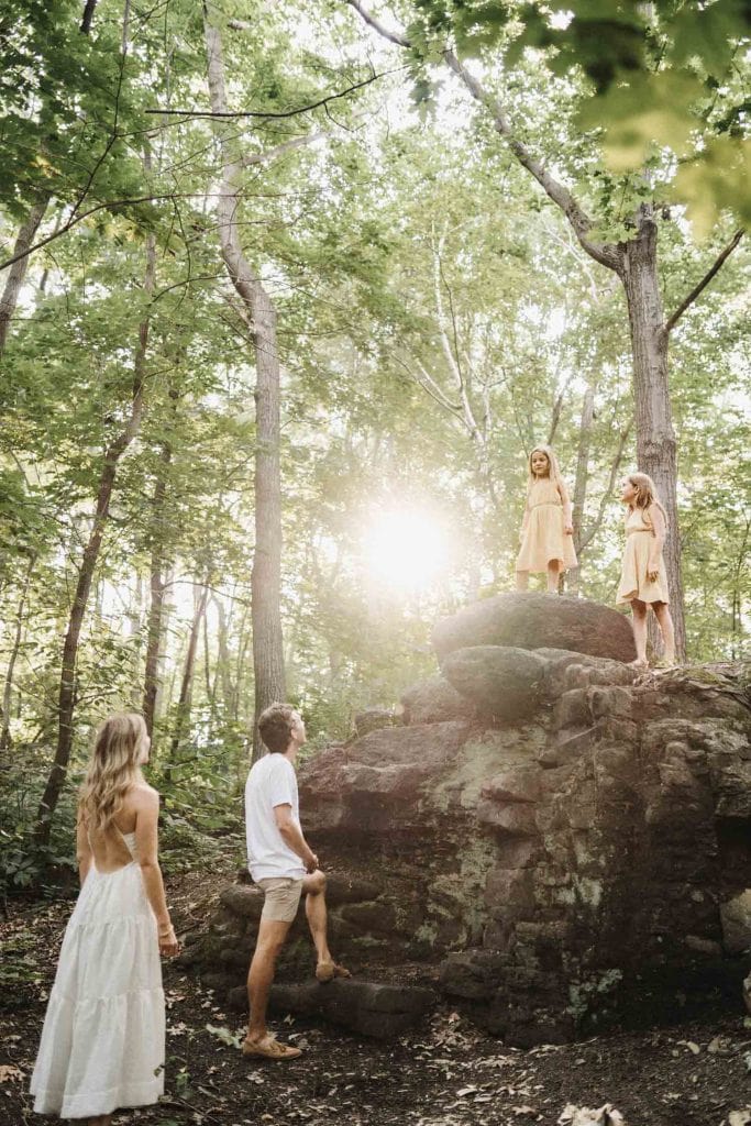 A family enjoying a sunny day in a lush Montreal forest, with two children standing atop a large rock, bathed in sunlight, as their parents watch admiringly from below. The children, dressed in matching yellow dresses, and the parents, in light summer attire, are surrounded by towering green trees, capturing a moment of adventure and family bonding