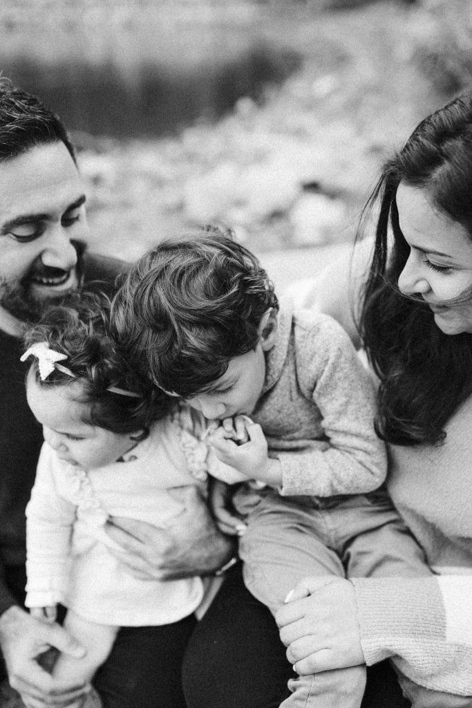 A black and white family portrait outdoors, showing parents and their two young children huddled close together, sharing a moment of quiet intimacy by a riverbank.