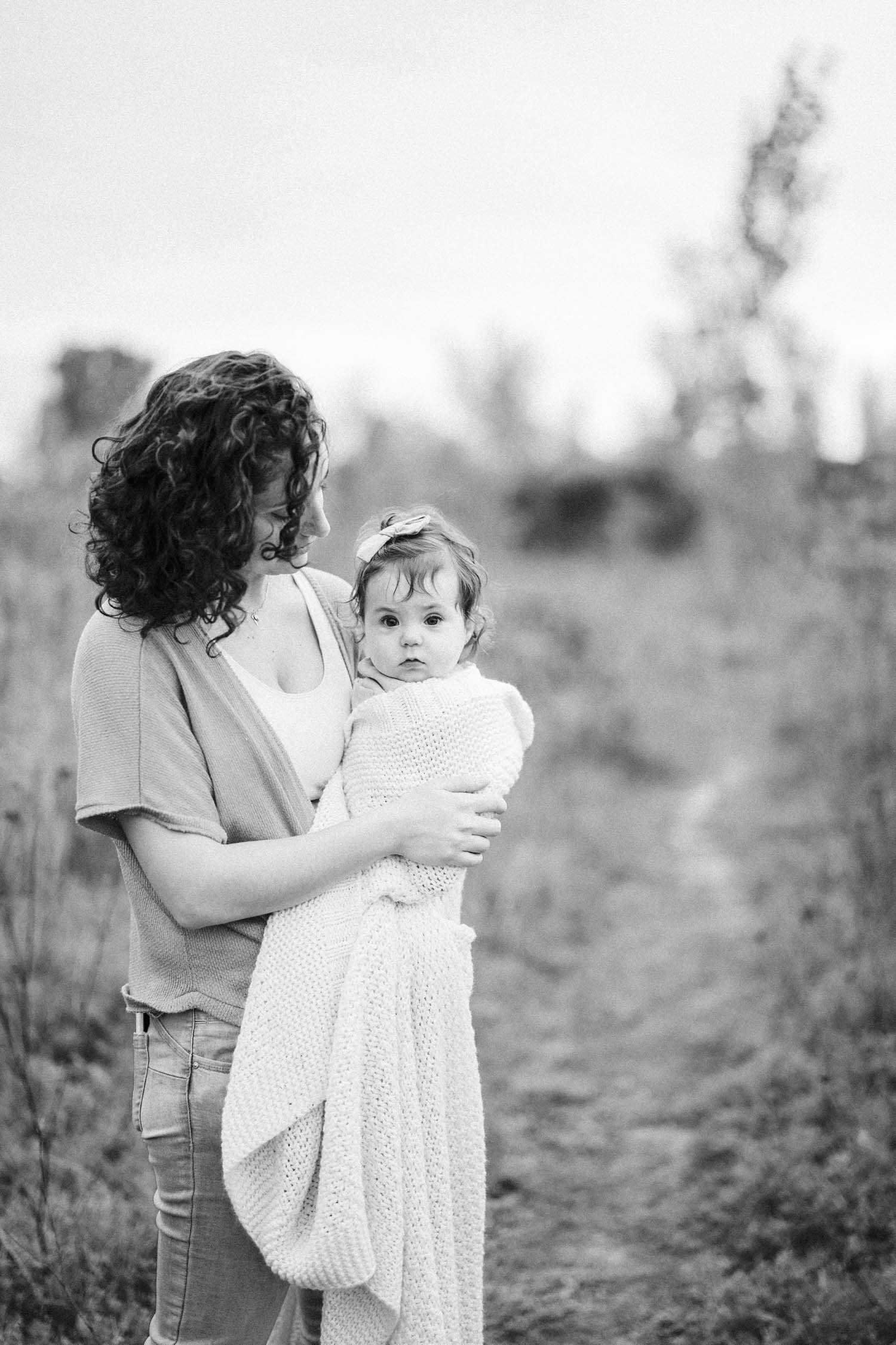 Mom holds her little girl close, while standing surrounding by greener during their photo session in Laval