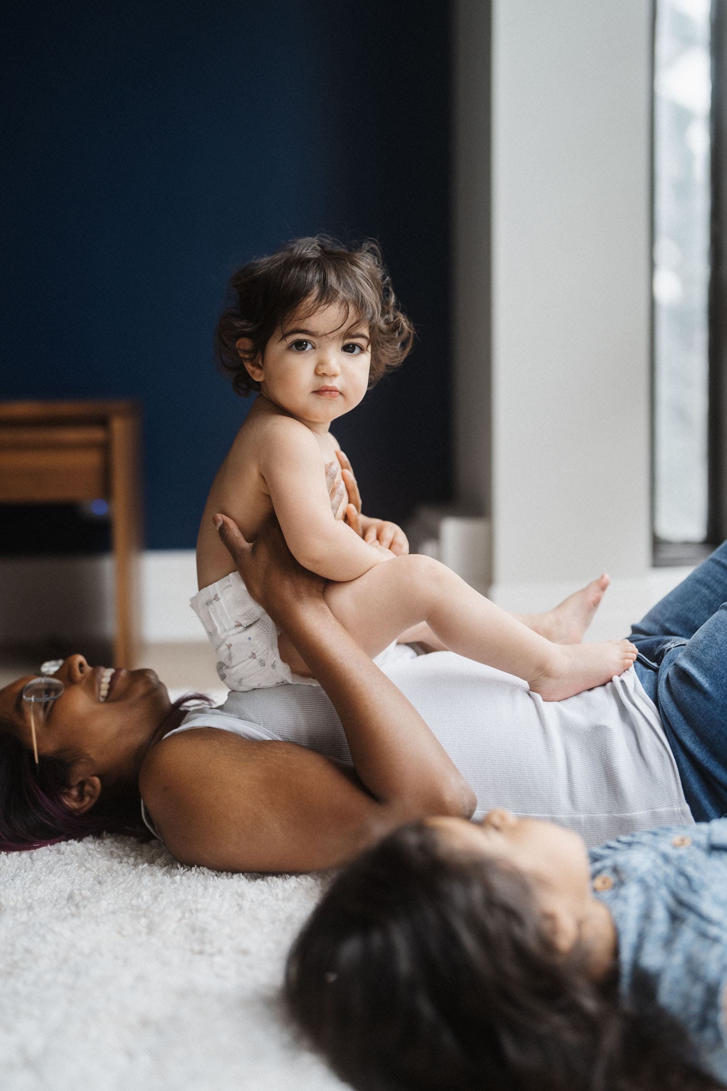 Baby in diaper sits on mom's stomach looking at camera, during their in-home motherhood family session in Montreal